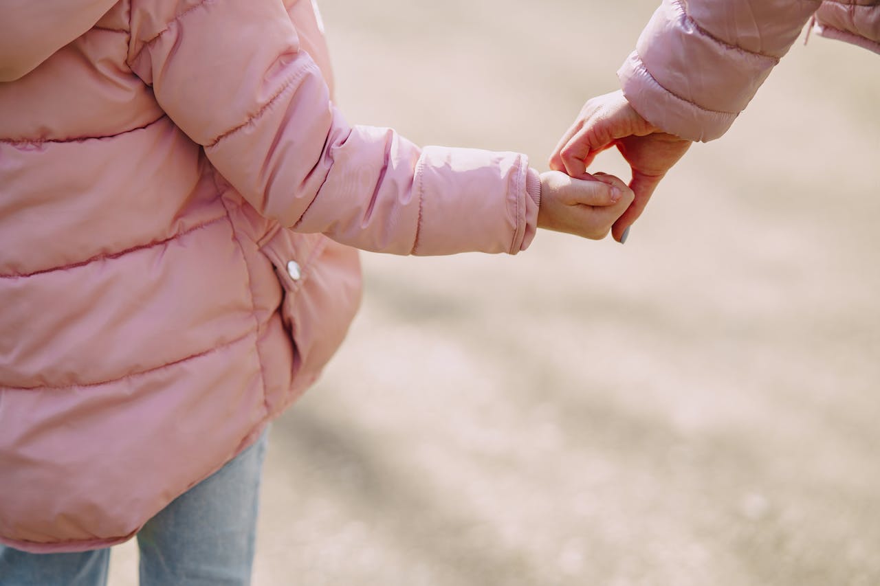 Back view of unrecognizable little girl wearing warm pink jacket holding hands with crop young woman while spending time together in city outside illustrating mother care and support concept